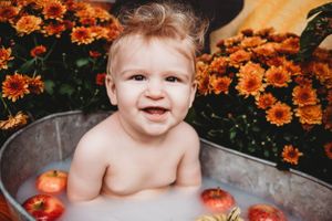 Smiling baby sits in metal tub surrounded by pumpkins and orange flowers.