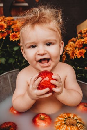 Happy baby in metal tub holding red apples among fall decorations and orange flowers.