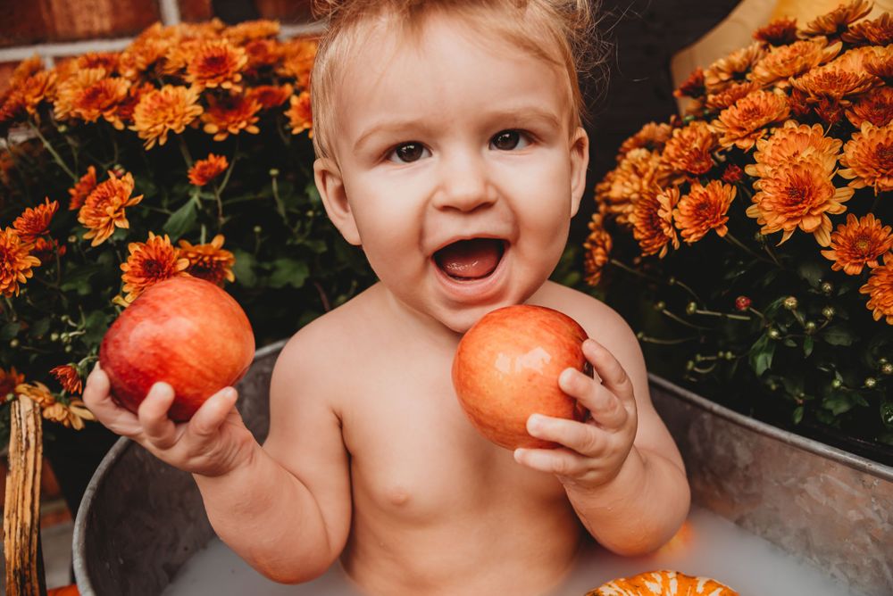 Joyful toddler holding oranges in a metal tub surrounded by bright fall mums.