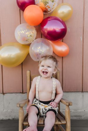 A happy child sits in a wooden chair with colorful balloons against a pink wall.