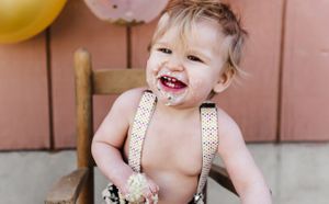 Happy toddler with messy face wearing suspenders and sitting in wooden chair.