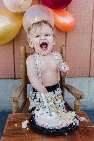 A messy and joyful cake smash photo with a laughing child surrounded by colorful balloons.