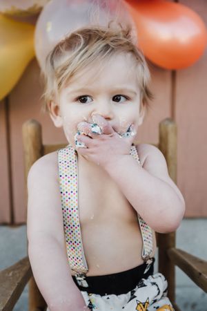 A child in polka dot suspenders giggles while covering their mouth sitting in front of balloons.