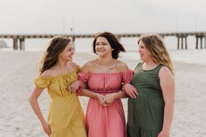 Three friends in summer dresses laughing together on a sandy beach with a pier in the background.