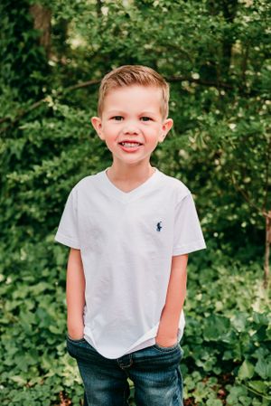Young child wearing a white polo smiling in front of green foliage.