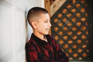Young person in burgundy velvet shirt smiles while sitting against white wall with decorative lattice background.