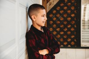 Young person in a red and black plaid shirt stands with arms crossed against white wall near decorative wooden door.