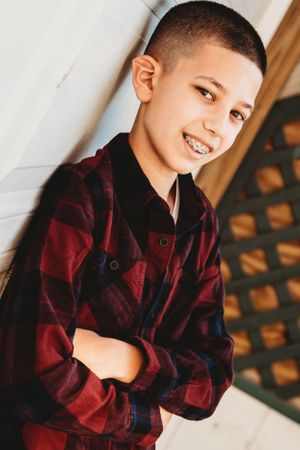 Young person in a red and black plaid flannel shirt poses near a wall with natural lighting.