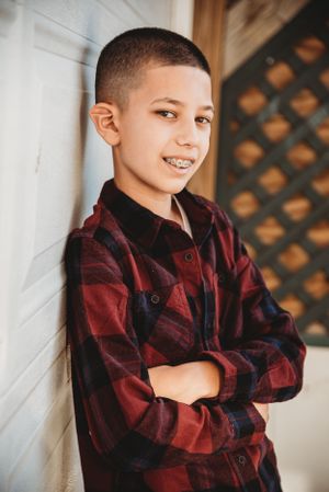 Young person in a red and black plaid flannel shirt leans against white wall with crossed arms.