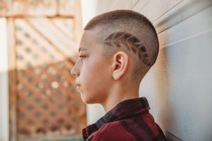 Side profile view of a young person with a stylish geometric hair design wearing a burgundy jacket against a light wall.