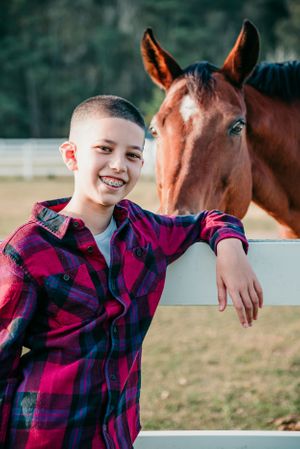 A friendly interaction between a young child in a red plaid shirt and a brown horse by a white fence.