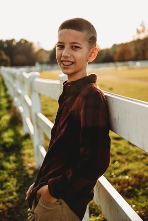 A child in a plaid shirt leans against a white fence on a sunny autumn day with golden light in the background.