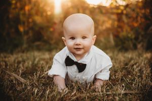 Baby wearing white dress shirt and bowtie crawls on grass during golden sunset.