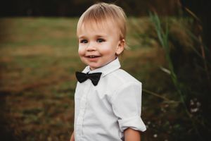 A toddler wearing a white dress shirt and black bow tie smiles while standing outdoors near tall grass.