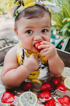 Baby wearing yellow floral outfit enjoying fresh strawberries and citrus fruits.