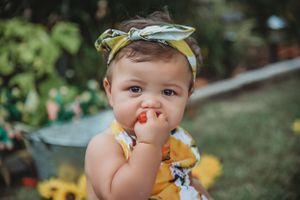 Baby in yellow floral outfit and matching headband eating fresh fruit outdoors.