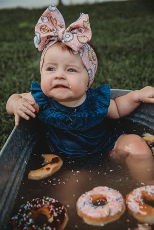 Baby in blue dress and floral headband sitting in basin with donuts.