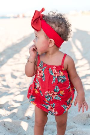 Small child in red floral romper and bow headband playing on sandy beach.