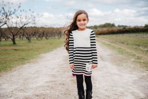 A young child in a striped dress walks on a dirt path through an orchard under cloudy skies.