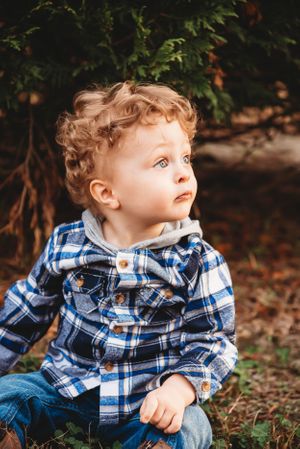 Small child wearing plaid blue and white flannel shirt sits outdoors near greenery in warm autumn lighting.