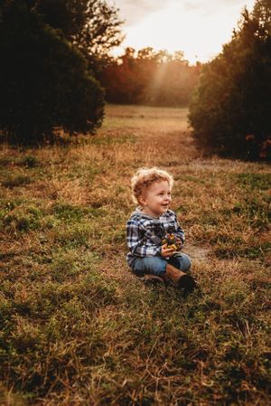 Child sits in grassy field wearing plaid shirt at golden hour sunset with trees in background.