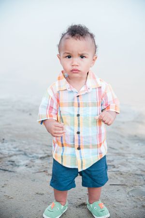 Young child in pastel plaid shirt and blue shorts standing on beach.