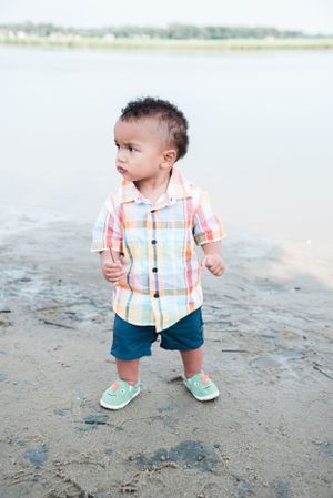 A toddler in plaid shirt and shorts standing on a sandy beach near calm water.