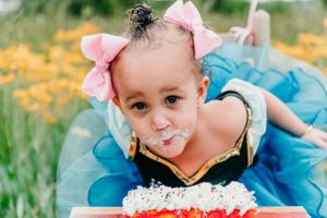 A small child in a blue dress with pink bows enjoying a frosted cake outdoors among yellow flowers.