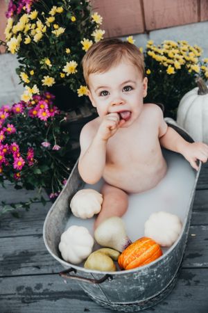 Baby sits in metal tub with pumpkins and gourds amid colorful fall flowers.