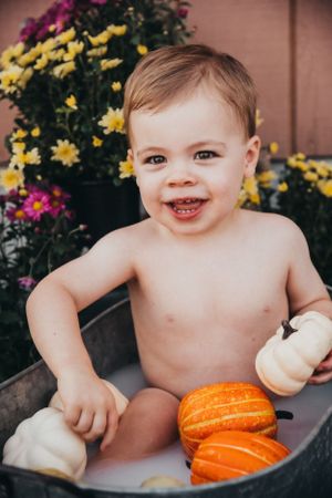 Smiling baby poses in vintage tub with white and orange pumpkins surrounded by flowers.