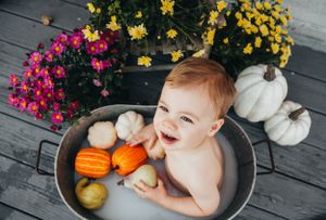 Baby laughs while sitting in metal tub with autumn gourds and colorful mums.