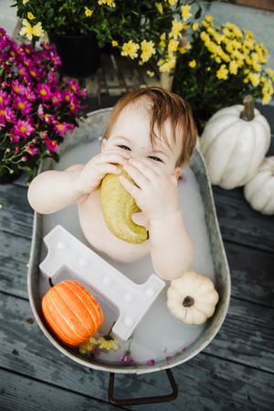 Baby in metal tub playfully bites pear among decorative pumpkins and flowers.