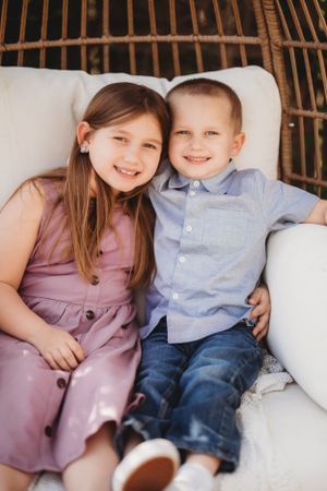 Two children in coordinating outfits smile while sitting together on white cushions.