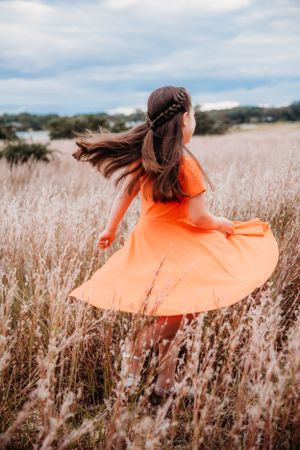 Young girl in flowing orange dress twirling joyfully in a field of tall grass under cloudy skies.