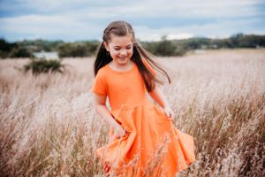 Girl in bright orange dress laughing while dancing in a field of dried grass.