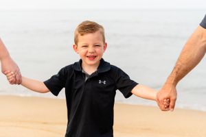 Child in black Under Armour shirt smiling and holding hands on sandy beach.