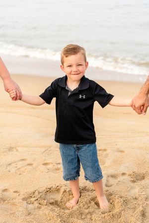 A young child in a black polo shirt and denim shorts holds hands with adults while standing barefoot on a sandy beach.