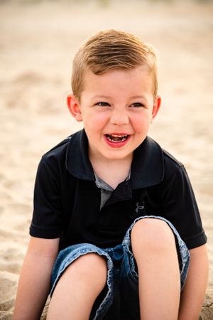 A joyful child wearing a black polo shirt and denim shorts sits on sandy ground in warm sunlight.