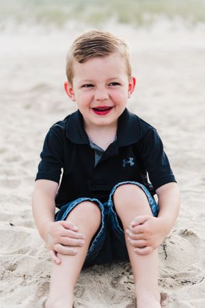 A child in a navy blue Under Armour polo sits smiling on a sandy beach.