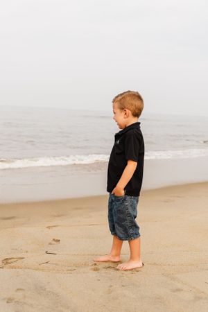 Young child in a black shirt and denim shorts stands barefoot on a misty beach looking at the ocean.