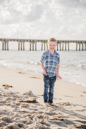 A plaid-shirted child standing on a beach with a wooden pier in the background on a cloudy day.