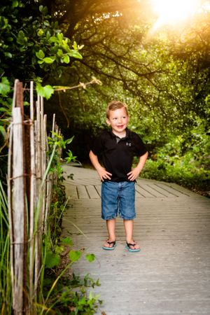 A child in a black polo and shorts stands on a sunlit wooden boardwalk path surrounded by lush green foliage.