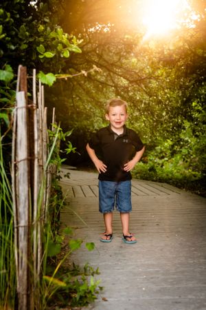 A child stands on a wooden boardwalk path through green foliage at sunset with golden sun flare in background.