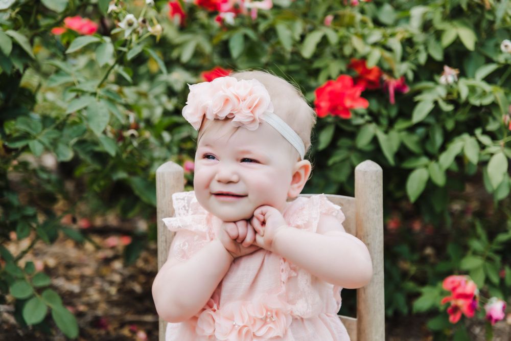 Smiling baby in pink dress sits in rustic wooden chair near blooming garden.