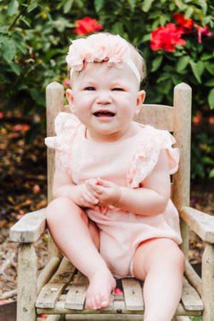 Baby wearing pink outfit and headband sits in wooden chair with red flowers in background.