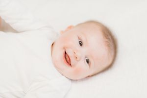 A smiling baby in white clothing lies on a light colored background.