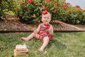 Baby in watermelon print outfit sitting outdoors with cake slice near red flowers.