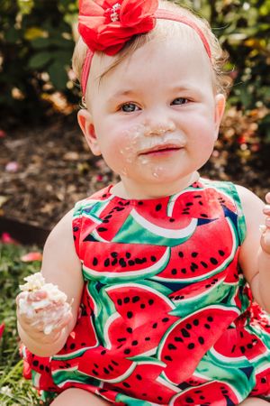 Close up of baby in watermelon print outfit enjoying cake during outdoor photoshoot.