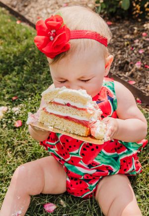 Baby wearing watermelon print outfit taking big bite of layered cake in garden setting.