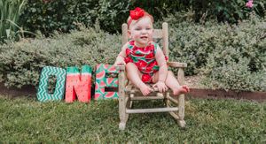 Baby in watermelon romper sitting on wooden chair next to decorative ONE letters outdoors.